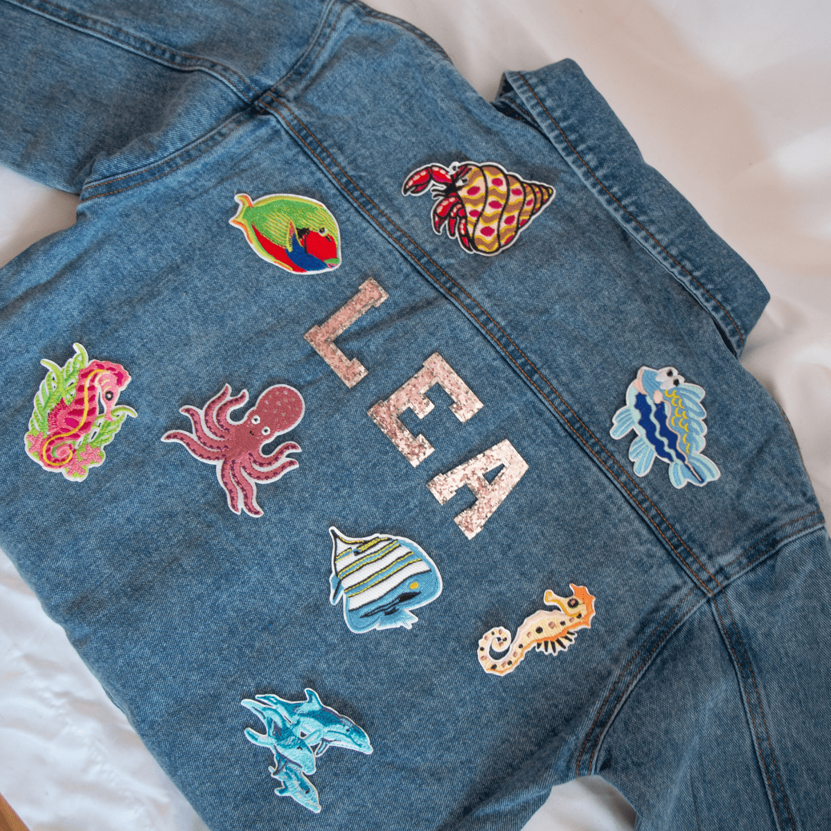 Dunkelblaue Jeansjacke für Kinder mit glitzernden Buchstaben und bunten Meerestieren-Bügelbildern – ein kreatives und maritimes Statement-Piece für kleine Abenteurer.